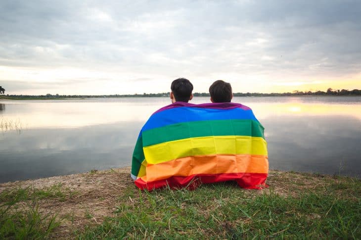 Two young man covered by a rainbow flag  beside the lake on sunset sky background. LGBT, Gay, Pride Concept.